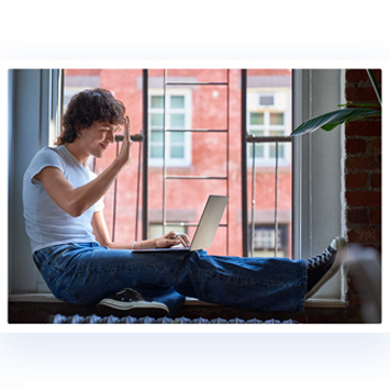 Eine Person sitzt am Fenster mit Laptop, trägt Jeans und ein weißes T-Shirt, Gebäude im Hintergrund.