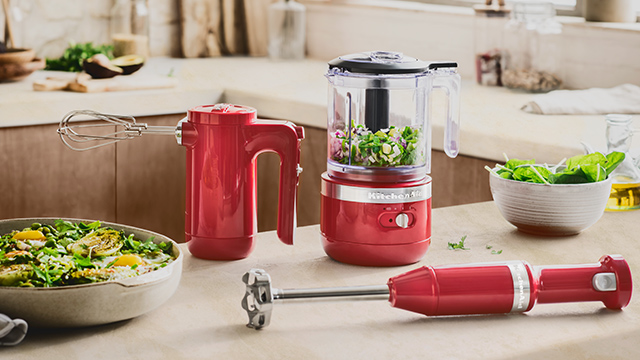 Küchenszene mit rotem Stabmixer, Handmixer mit Rühraufsatz, Zerkleinerer mit Gemüse, Salatschüssel und Zutaten auf einer Arbeitsfläche.