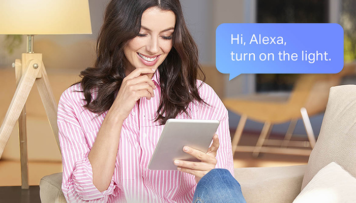 Eine Frau sitzt auf einem Sessel und hält ein Tablet. Über ihr befindet sich eine Sprechblase mit der Aufschrift "Hi, Alexa, schalte das Licht ein".