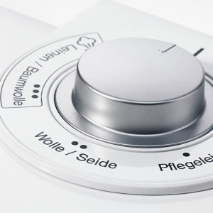 Nahaufnahme eines weißen Waschmaschinendrehknopfes mit silberfarbenem Akzent und Programmen wie "Wolle/Seide" und "Pflegeleicht".