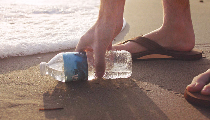 Eine Hand greift nach einer Plastikwasserflasche am Strand. Im Hintergrund sind Füße in Sandalen und Wellen zu sehen.