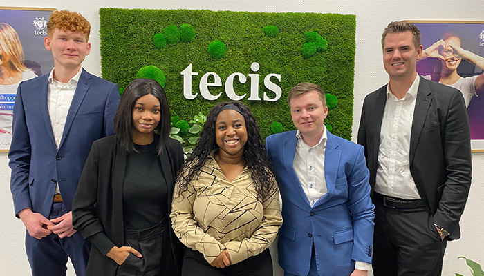 Fünf Personen in Anzügen stehen vor einer grünen Wand mit dem weißen "tecis"-Logo, eingerahmt von Postern.