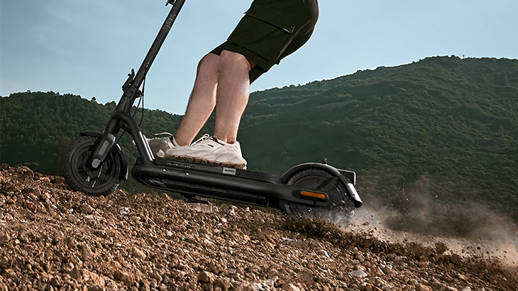 Eine Person fährt mit einem schwarzen Elektroroller auf einem unbefestigten Weg, mit einem bewaldeten Hügel im Hintergrund.