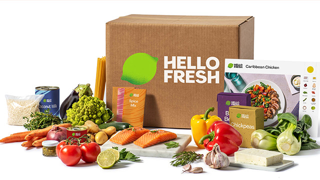 Eine Hello Fresh Box mit Gemüse, Pasta, Fisch und Kochzutaten auf einem weißen Tisch vor weißem Hintergrund.