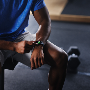 Person im Fitnessstudio, die auf einer Bank sitzt und ihre Sport-Smartwatch am Handgelenk bedient, im Hintergrund liegen Hanteln auf dem Boden.