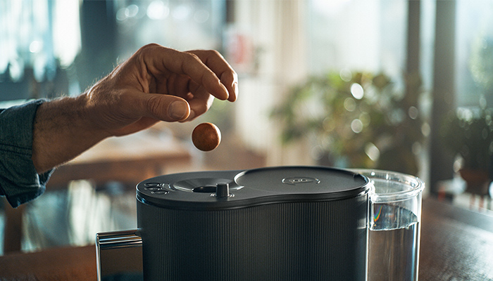 Eine Hand lässt eine Kaffeekapsel über eine Kaffeemaschine fallen. Daneben steht ein Wasserbehälter.