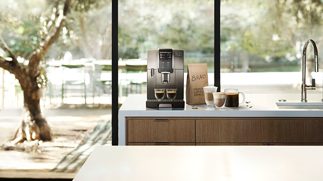 Auf einer Küchenarbeitsplatte stehen eine Kaffeemaschine, zwei Tassen Kaffee, eine Kaffeebohne und eine Kaffeetüte vor einem Fenster.