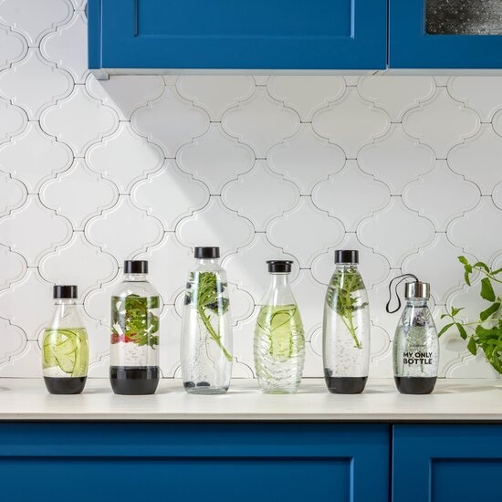 Sechs Wasserflaschen stehen auf einer Theke vor blauen Schränken und weißen Fliesen. Einige Flaschen enthalten Gurken und Kräuter.