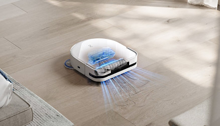 Ein weißer Saugroboter mit blauen Lichteffekten reinigt einen hellen Holzboden. Teile eines Sofas sind im Bildvordergrund sichtbar.