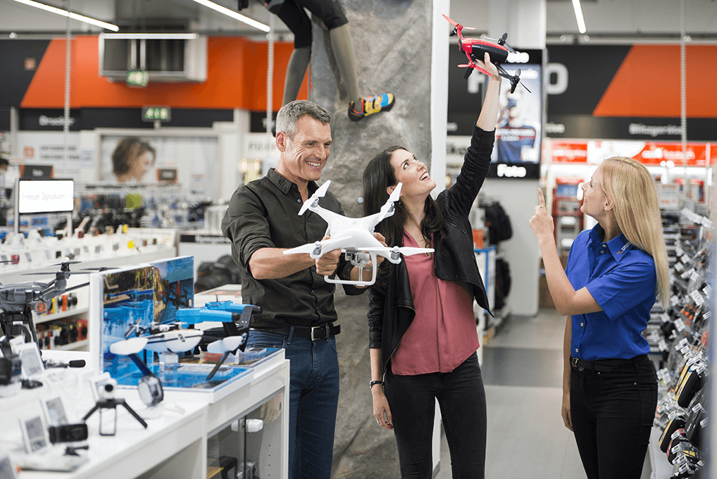 In einem Geschäft hält ein Mann eine weisse Drohne, während eine Frau eine rot-schwarze Drohne hochhält. Eine Verkäuferin zeigt.