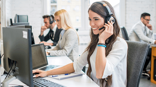 In einem Büro sitzen mehrere Personen mit Headsets an Computern. Eine Frau lächelt und telefoniert.