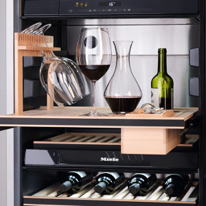 Im Weinkühlschrank stehen Weingläser, eine Karaffe und eine grüne Weinflasche auf einem Holzregal über mehreren Flaschen Wein.