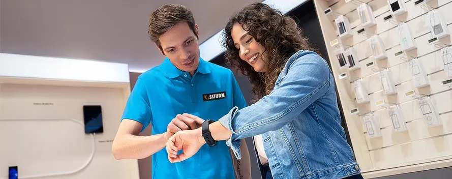 In einem Geschäft probiert eine Frau eine Smartwatch an, während ein Verkäufer in blauem Hemd zusieht.
