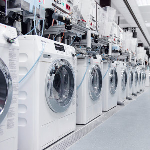 Eine Reihe weißer Waschmaschinen mit silbernen Türen stehen in einer hellen Halle unter industriellen Anlagen.