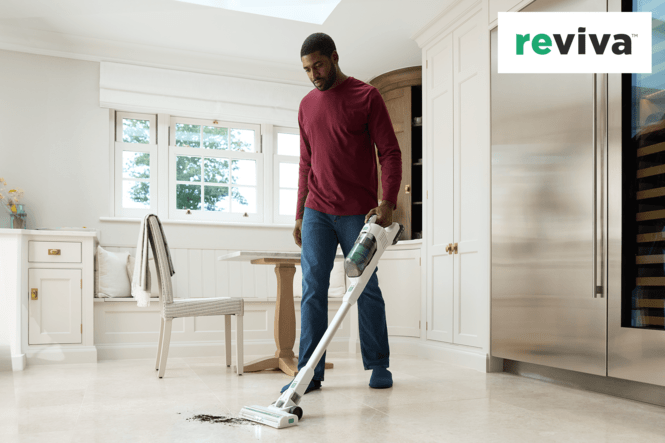 Ein Mann saugt mit einem weißen Staubsauger in einem hellen Raum. Im Hintergrund befinden sich Fenster, ein Tisch und Küchenschränke.