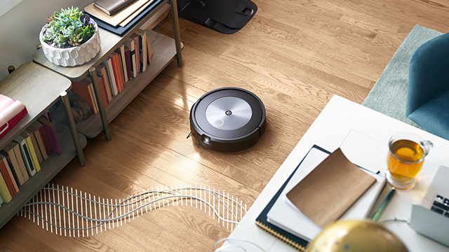 Ein runder, schwarzer Staubsaugerroboter steht auf einem Holzboden neben einem Tisch und einem Bücherregal.