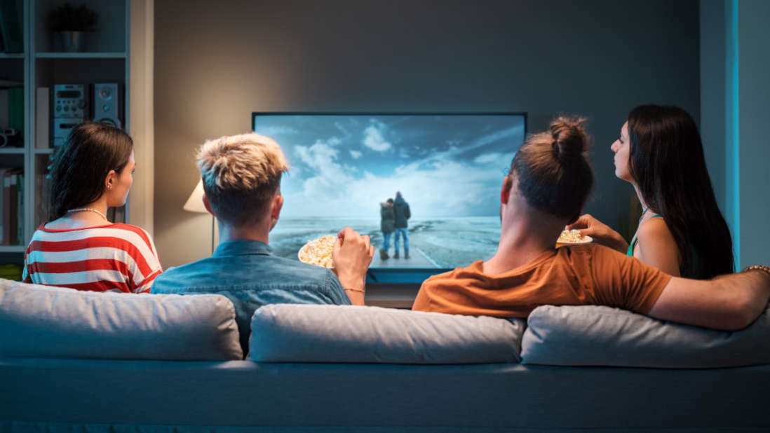 Vier Personen sitzen auf einem Sofa und schauen einen Film auf einem großen Fernsehbildschirm.