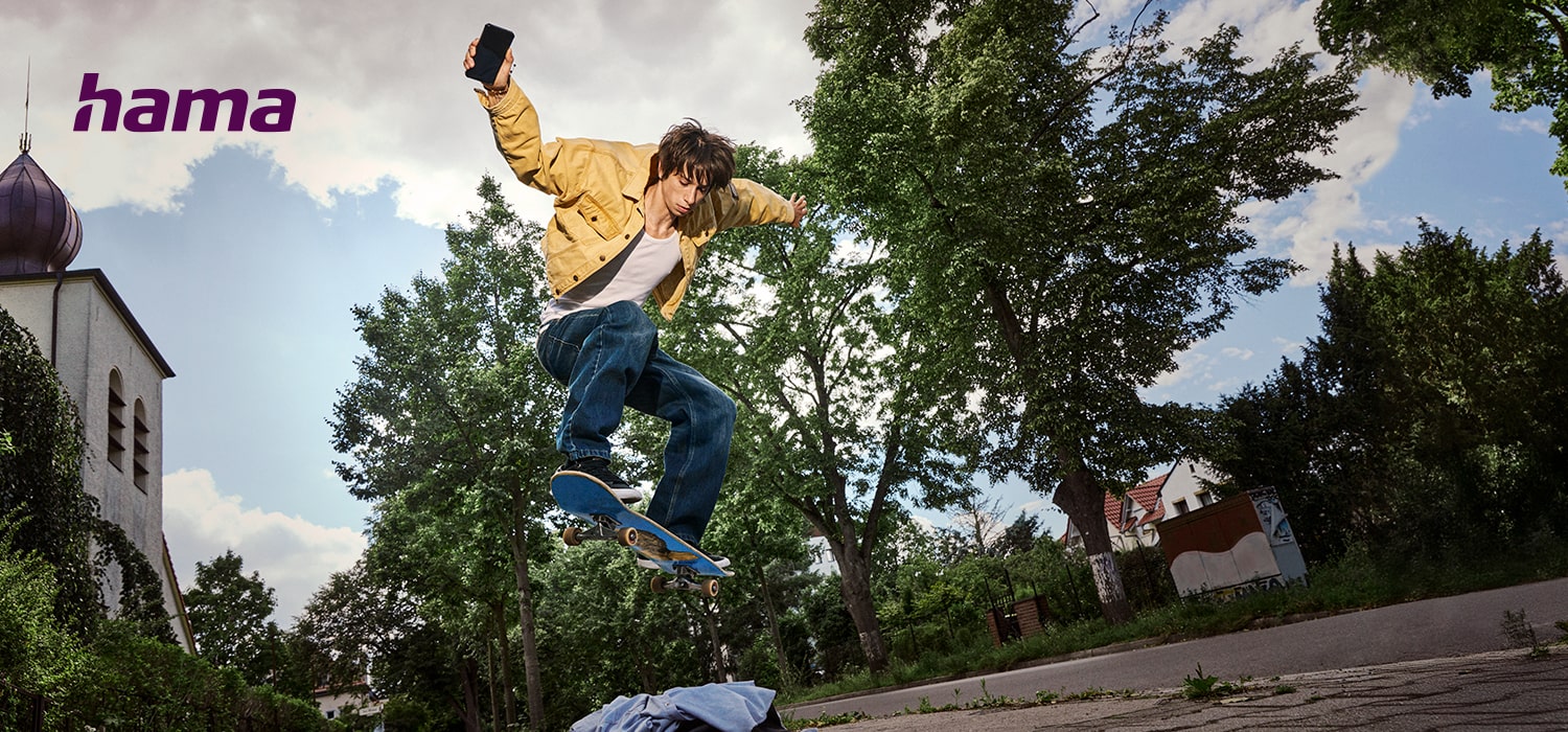 Skateboarder mit Smartphone in der Hand springt auf der Straße mit seinem Board in die Luft, im Hintergrund Bäume und Gebäude, „hama“-Logo sichtbar.