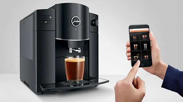 Schwarzer Kaffeevollautomat mit Glas Kaffee darunter, bedient per Handy, App zeigt Kaffeeauswahl.