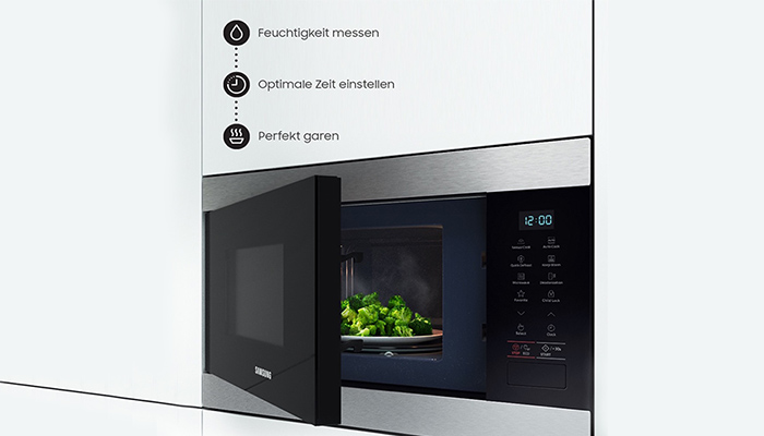 Eine Mikrowelle mit teilweise geöffneter Tür zeigt Gemüse auf einem Teller im Inneren. Die Anzeige zeigt 12:00.