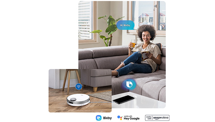 Eine Frau liest auf einem grauen Sofa, während ein Saugroboter auf dem Holzboden reinigt. Ein Smartphone liegt auf einem Tisch.