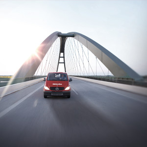 Roter Transporter fährt auf einer Brücke mit Stahlseilen vor hellem Himmel und Sonnenlicht.