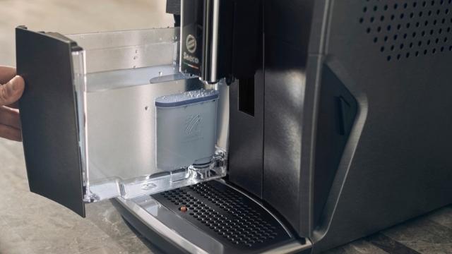 Eine Hand öffnet die Tür einer schwarzen Kaffeemaschine, die einen silbernen Wassertank mit blauem Filter zeigt.