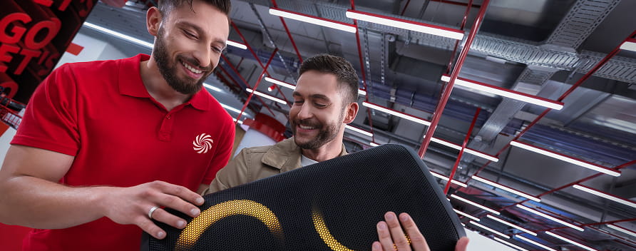 Verkäufer im roten Poloshirt zeigt einem Kunden lächelnd einen schwarzen Lautsprecher im Elektronikmarkt.