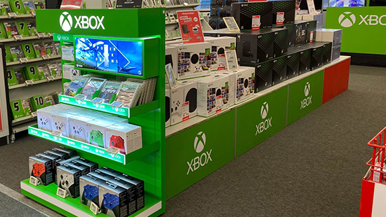 Grüne Xbox-Verkaufsstände mit Konsolen, Spielen und Zubehör in einem Geschäft. Eine Produktanzeige zeigt Bildschirme und Packungen.
