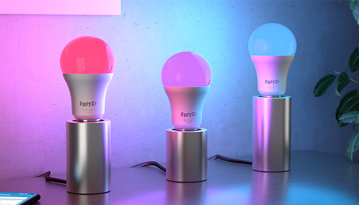 Drei FRITZ!-Glühbirnen mit silbernen Sockeln leuchten in Rot, Lila und Blau auf einem Tisch vor einer blauen Wand mit Pflanze.