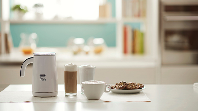 Ein weißer Milchaufschäumer steht neben einem Glas Latte Macchiato, einer Tasse und einem Teller Kekse auf einem Tisch.