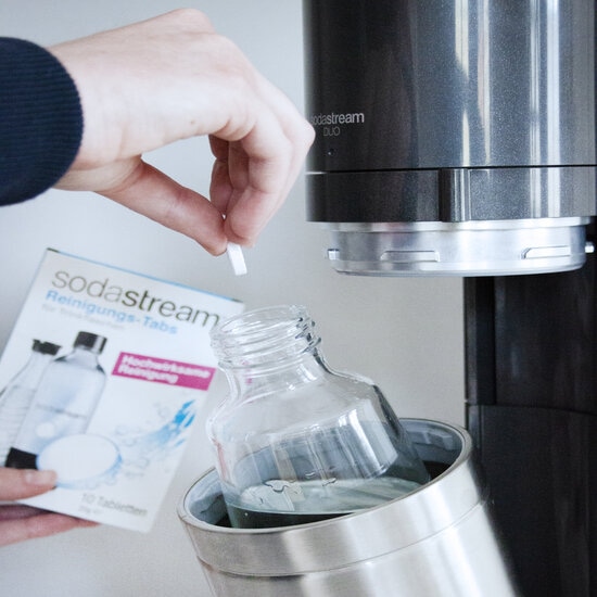 Eine Person gibt eine Reinigungstablette in eine Glasflasche, die in einem SodaStream-Gerät steht.