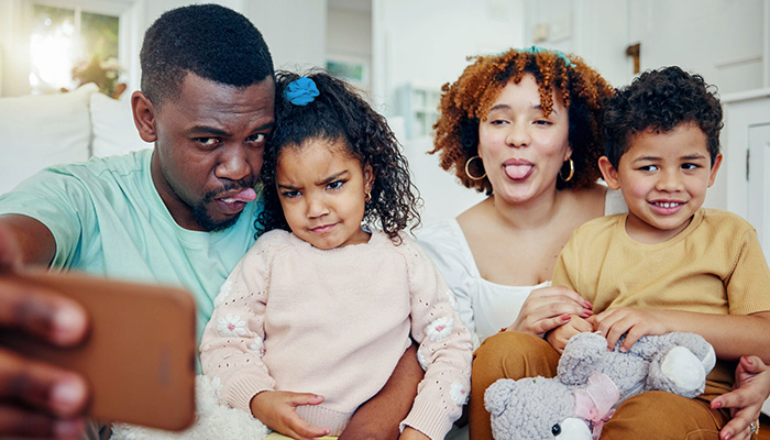 Eine Familie macht ein Selfie, bei dem die Eltern und Kinder alberne Gesichter ziehen. Der Junge hält einen Teddybären.