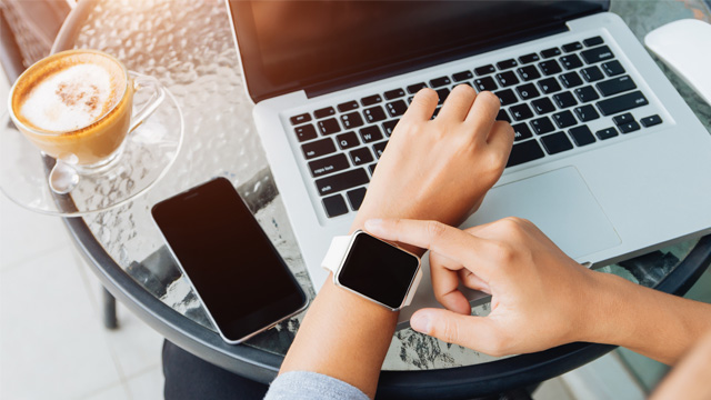Eine Person tippt auf einem Laptop mit einer Smartwatch am Handgelenk, neben einem Smartphone und einer Tasse Kaffee auf einem Tisch.