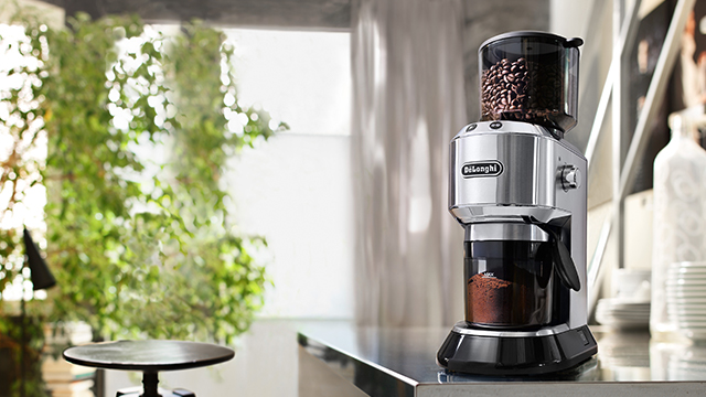 Ein silberner DeLonghi-Kaffeemühle steht auf einer glänzenden Theke, mit Bohnen oben und Kaffeepulver unten. Im Hintergrund Fenster mit Grün.