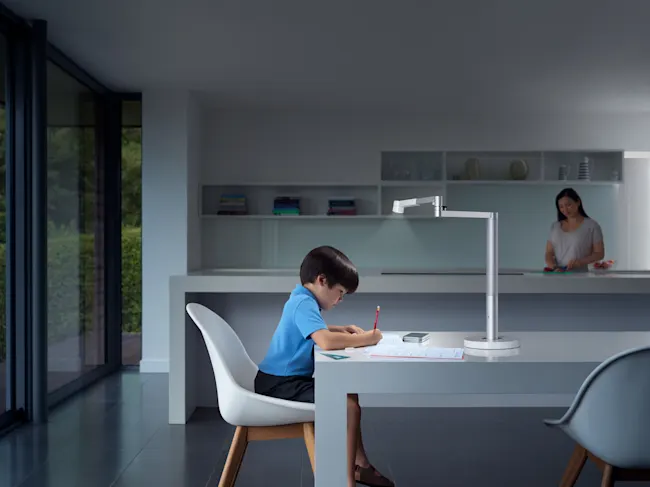 Ein Kind sitzt an einem Tisch und schreibt, beleuchtet von einer Lampe. Im Hintergrund steht eine Frau in einer Küche.