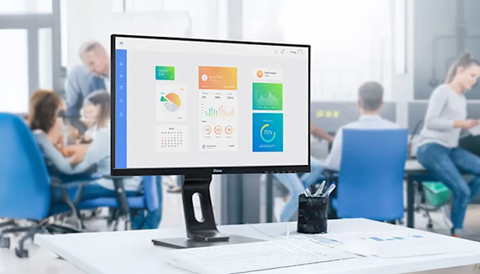 Ein Computerbildschirm zeigt Diagramme und Kalender. Ein Stifthalter steht auf einem weißen Tisch. Im Hintergrund sitzen Personen.