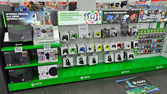 Ein Verkaufsstand mit Xbox-Konsolen, Controllern und Zubehör auf grünen Regalen in einem Geschäft. Werbeschilder sind auch sichtbar.