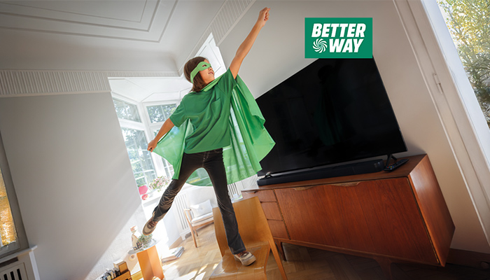 Ein Kind mit grüner Maske und Umhang steht auf einem Stuhl vor einem großen Fernseher in einem hellen Raum.