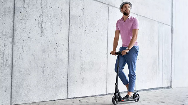 Mann mit Hut und rosa Hemd auf einem Roller vor grauer Wand.