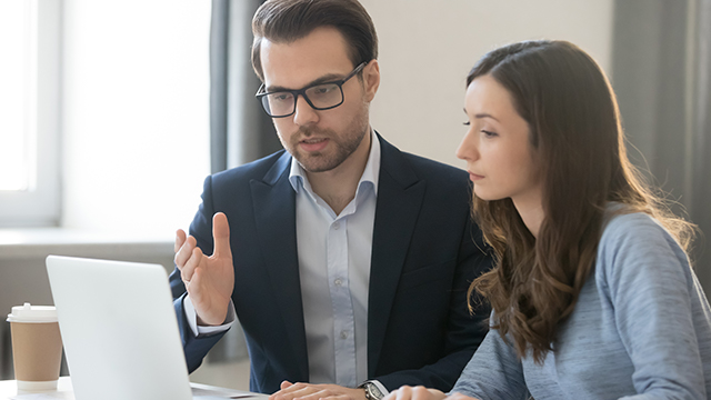 Mann im Anzug gestikuliert vor Laptop, Frau hört zu. Kaffeetasse und Fenster im Hintergrund.