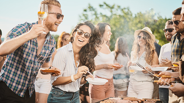 Eine Gruppe von Leuten grillt im Freien, einige halten Teller und Gabeln, während andere Getränke halten.