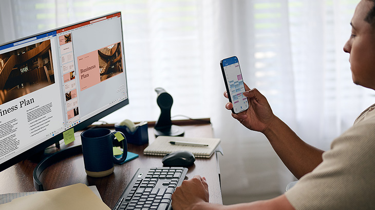 Ein Mann sitzt mit einem Smartphone im Hand an einem Arbeitstisch. Die Softwares Word, PowerPoint und Outlook sind sichtbar.