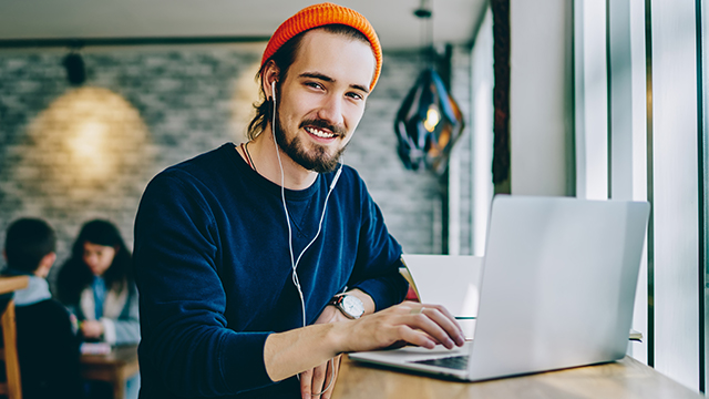 Ein Mann mit orangefarbener Mütze tippt auf einem Laptop, trägt Kopfhörer und eine dunkelblaue Bluse in einem Café.