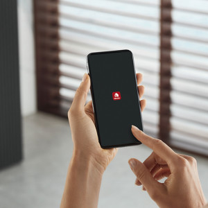 Eine Hand hält ein schwarzes Smartphone mit einem roten Symbol vor einem Fenster mit Jalousien.