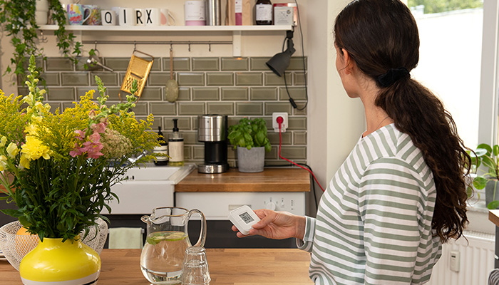 Eine Frau in gestreiftem Oberteil hält eine Smart Home Fernbedienung in der Hand in einer Küche mit Blumen und Küchengeräten.