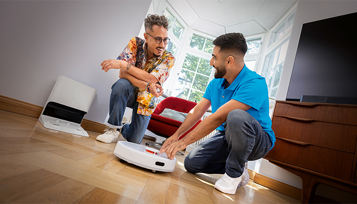 Zwei Männer hocken auf Holzboden bei einem Roboterstaubsauger, in einem Raum mit Fenster, Kommode und Ladestation.