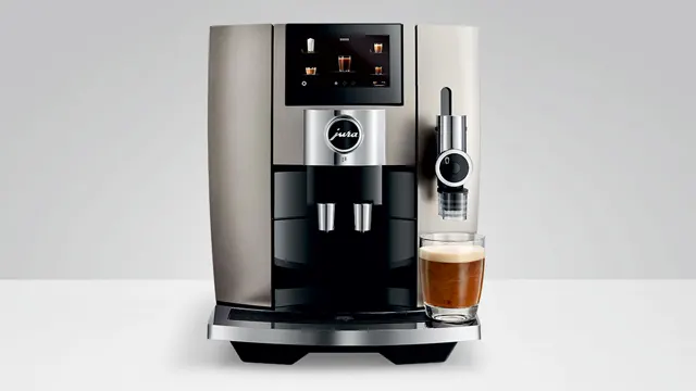 Ein Kaffeevollautomat mit einem Glas Kaffee auf einer hellen Oberfläche vor einem neutralen Hintergrund.