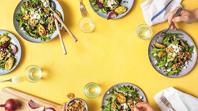 Auf gelbem Grund sind Salatteller, Getränke, Besteck, Walnüsse und Zwiebeln angeordnet. Hände halten Gabeln.