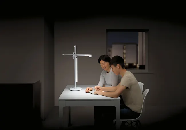 Ein Mann und eine Frau sitzen an einem weißen Tisch unter einer Lampe und schreiben in ein Notizbuch. Im Hintergrund ist ein Fenster.